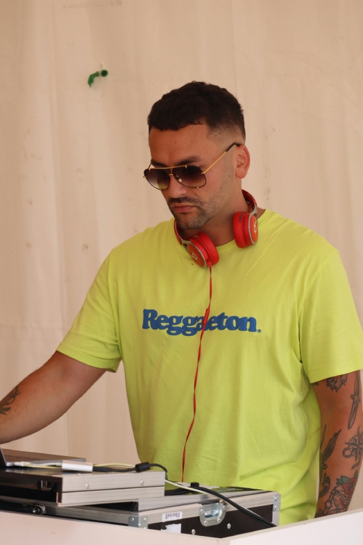 Man DJ'ing wearing a sunglasses and a yellow top reading reggaeton.
